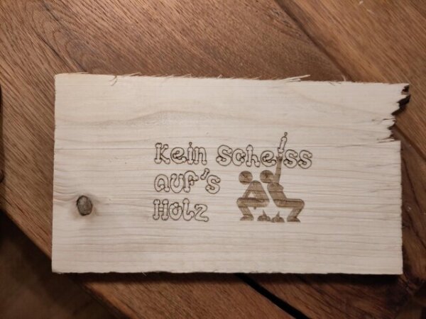 Holzschild mit gelaserter Aufschrift "Kein Scheiss auf's Holz" und zwei kackenden Menschen.